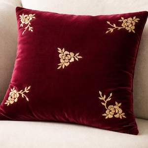 Pode incluir: Uma almofada quadrada de veludo bordô com bordado floral dourado. As flores bordadas são colocadas em cada canto e no centro da almofada. A almofada está em um sofá de cor clara.