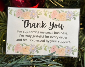 以下が含まれることがあります： 「Thank you」の文字が黒い筆記体で書かれた白いカード。ピンク、黄色、紫の花のイラストで囲まれています。カードのメッセージは、小さなビジネスをサポートしてくれたことへの感謝を表しており、クリスマスツリーと赤いオーナメントを背景にしています。