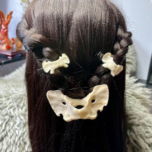 May include: Dark brown hair styled in braids, adorned with three bone-colored hair clips. The clips feature a unique, organic design, adding a touch of natural elegance to the hairstyle. The clips are secured with black metal clasps.