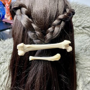 May include: Two bone-shaped hair clips are placed in dark brown hair styled in a braid. The clips are off-white and appear to be made of bone. The hair is long and flowing, with a slight wave.