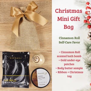 May include: A Christmas mini gift bag with a gold ribbon bow, containing a cinnamon roll scented bath bomb, gold under-eye patches, and body butter. The bag is on a wooden surface. Text on the image reads "Christmas Mini Gift Bag".