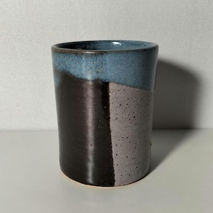 May include: A handcrafted ceramic tumbler with a distinctive colour-blocked design. The top is a muted blue, transitioning to a dark brown and a speckled grey. The cylindrical cup is suitable for beverages.