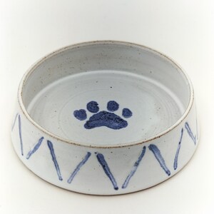 May include: A round, off-white ceramic pet bowl with a speckled finish. The interior base features a single blue paw print design. The exterior side of the bowl is decorated with a repeating pattern of vertical, hand-painted blue brush strokes.