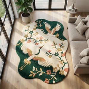 Puede incluir: Una alfombra de forma irregular con un centro de color crema que presenta dos grullas volando y ramas florecientes en tonos rosa, amarillo y verde. La alfombra tiene un borde verde oscuro e incluye el texto Tufting Carpets en el centro.