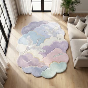 Op de afbeelding: Een wolk-vormig vloerkleed in pastelkleuren van lavendel, blauw, roze en mintgroen. Twee witte kraanvogels zijn afgebeeld in vlucht. Het kleed heeft een zachte, pluche textuur en ligt op een houten vloer.