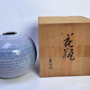 May include: A spherical ceramic vase with a mottled light blue and grey glaze, featuring a small circular opening at the top. Beside it sits a square wooden storage box with black Japanese calligraphy characters printed on the front surface.
