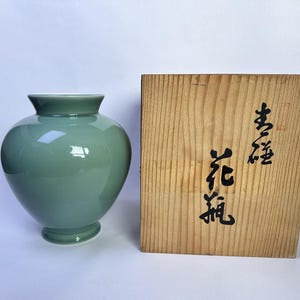 May include: A smooth, sage green ceramic vase with a rounded body and a flared rim, positioned next to a rectangular wooden storage box featuring black Japanese calligraphy characters printed vertically on the front surface of the light wood.