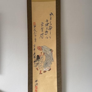 May include: A vertical hanging scroll featuring a painting of a bald figure in a light blue robe interacting with a child in a striped garment. The artwork includes black Japanese calligraphy, red seal stamps, and a dark brown fabric border frame.