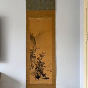 May include: A vertical hanging scroll featuring a monochromatic ink wash painting on a tan background. The artwork depicts a landscape with a waterfall, rocky cliffs, and pine trees. The scroll has dark green fabric borders at the top and bottom.