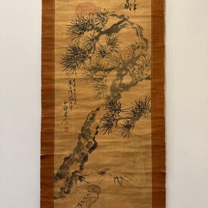 Op de afbeelding: Een verticale hangende rol met een schildering op beige papier, voorzien van een zwarte inktillustratie van een dennenboom en een rode zon. Het kunstwerk bevat zwarte kalligrafische tekst en een klein rood zegel. De rol heeft bruine stoffen randen.