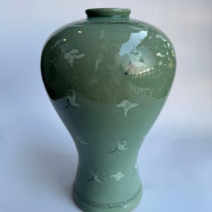 May include: A sage green ceramic vase with a glossy crackle glaze finish. The vase features a rounded body that tapers toward the base and neck, decorated with small, white, stylized bird motifs scattered across the surface and linear patterns.