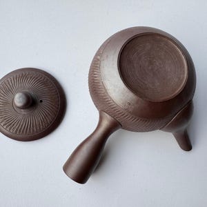 May include: A dark brown clay teapot with a side handle and a separate circular lid. The body and lid feature a radial, sunburst-like carved texture. A small, square-shaped maker's mark is visible on the side of the teapot body near the handle.