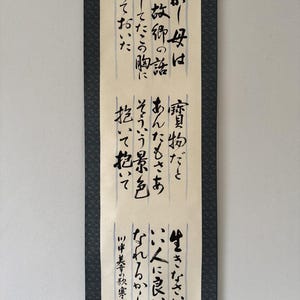 May include: A vertical hanging scroll featuring black Japanese calligraphy on a cream-coloured paper background. The scroll is framed by dark patterned fabric borders. The text is arranged in three distinct sections, each containing vertical lines of handwritten characters.
