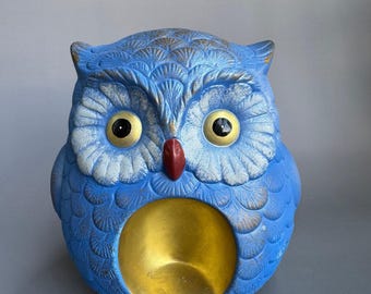 May include: A blue owl figurine with textured feather details, large yellow eyes with black pupils, and a dark red beak. The owl features a prominent, smooth, circular gold-colored hollow cavity on its chest and small clawed feet at the base.