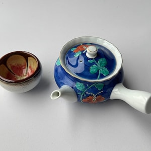 Könnte beinhalten: Eine Keramik-Teekanne mit seitlichem Griff und passendem Deckel, die eine tiefblaue Glasur mit orangefarbenen Blumen und grünen Blättern aufweist. Daneben steht eine kleine, runde Keramiktasse mit weißer Außenseite und rot bemaltem Blumenmuster innen.
