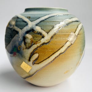 May include: A round ceramic vase featuring a gradient of blue, green, and tan tones with a textured surface. The exterior is decorated with white, raised, branching lines and small, square, metallic gold leaf accents applied to the glaze.