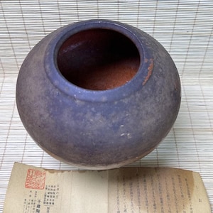 以下が含まれることがあります： 広い開口部と赤茶色の内部を持つ、丸い濃い紫色の陶器の壺。壺には、日本語のテキストと赤いスタンプが押された折りたたまれた紙の文書が添えられており、陶器の起源と工芸に関する情報が記載されています。