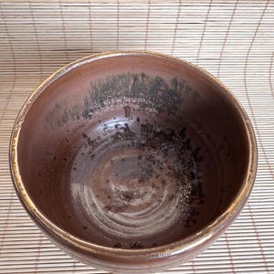 Może przedstawiać: Okrągła ceramiczna miska z ciemnobrązowym szkliwem, posiadająca nakrapiane, ciemne i teksturowane wnętrze. Krawędź ma jaśniejszy, przetarty wygląd, a cała powierzchnia wykazuje widoczne różnice w odcieniach i kolorystyce.