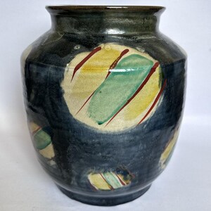 May include: A ceramic vase with a dark charcoal and black glazed finish. The surface features several circular motifs in cream, each decorated with diagonal stripes of sage green and deep red. The vase has a wide, rounded body and a short neck.