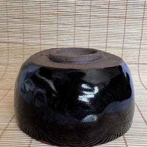 May include: A dark, glossy black ceramic bowl with an uneven, hand-formed surface texture. The bowl is inverted, revealing a thick, unglazed, dark brown circular foot ring at the top center. The finish is reflective, showing light highlights.