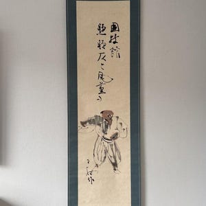 Op de afbeelding: Een verticale hangende rol met een beige papieren paneel gemonteerd op een donkerblauwe stoffen rand. De rol toont zwarte inktcalligrafie en een gestileerde inkttekening van een figuur in traditionele kleding met een bruine hoofdbedekking.