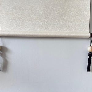 Könnte beinhalten: Eine gerollte Schriftrolle mit cremefarbenem Papier mit floralem Muster. Die Schriftrolle wird von dunkelbraunen Holzdübeln mit dekorativen Hängeperlen und schwarzen Quasten gehalten. Die Schriftrolle hängt an einer weißen Wand.