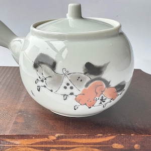 May include: White ceramic teapot with a lid and a side handle. The teapot is decorated with a hand-painted design of horses in black, gray, and orange. The teapot is sitting on a wooden surface.
