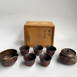 Pode incluir: Um conjunto de chá japonês com cinco pequenas chávenas de cerâmica castanho-escura, um bule a combinar com tampa e uma taça de servir. O conjunto é apresentado com uma caixa de arrumação retangular de madeira com caligrafia japonesa preta.