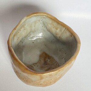 May include: A small, hand-formed ceramic bowl with an irregular, organic shape. The exterior features a textured, speckled tan and beige glaze, while the interior has a smooth, off-white finish with a prominent brown patch at the base.