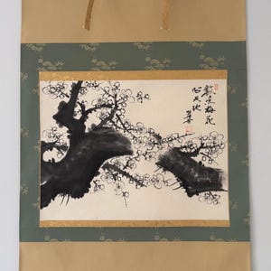 May include: A hanging wall scroll featuring an ink wash painting of a plum blossom tree branch with black ink strokes and delicate white flowers. The artwork includes vertical black calligraphy text and red stamps, mounted on a green and tan fabric border.