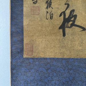 Könnte beinhalten: Eine vertikale Wandrolle aus gealtertem, beigefarbenem Papier mit schwarzer japanischer Kalligrafie. Das zentrale Papier ist von einem dunkelblauen Stoffrahmen mit wiederkehrendem Blumenmuster umgeben und enthält zwei rote Stempel.