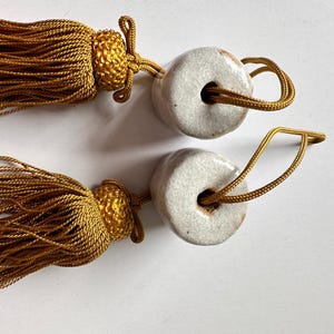 Puede incluir: Dos borlas decorativas con cordón dorado y borla dorada. Cada borla tiene un pequeño anillo de cerámica blanquecina. Las borlas están dispuestas sobre una superficie blanca.