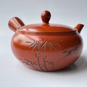 Puede incluir: Una tetera de cerámica de color marrón rojizo con un asa lateral y una tapa a juego con pomo. El cuerpo de la tetera está decorado con un diseño de bambú grabado que rodea la parte frontal, mostrando tallos y hojas detallados en marrón oscuro.