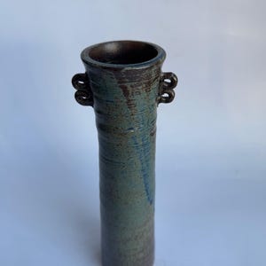 以下が含まれることがあります： 縁が広がった背の高い円筒形の陶器製花瓶で、両側に小さなループ状の取っ手がついています。表面は深い青、ティール、ダークブラウンの色合いのまだら模様の釉薬で覆われており、本体には水平方向の質感の線が見られます。