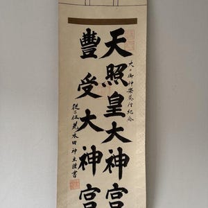 Op de afbeelding: Een verticale hangende rol met zwarte Japanse kalligrafie op een crèmekleurige stoffen achtergrond. De tekst bevat grote karakters die Ten-sho-ko-dai-jin-gu en Toyo-uke-dai-jin-gu lezen, met kleinere verticale tekst en rode zegels.