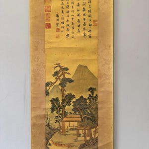 May include: A vertical hanging scroll painting on a gold-toned fabric mount. The artwork depicts a landscape with mountains, pine trees, and two small structures with figures inside. Black ink calligraphy and red seal stamps appear at the top.