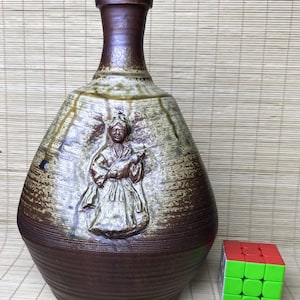 Puede incluir: Un jarrón de cerámica marrón y beige con cuello estrecho y cuerpo ancho y redondeado. La superficie presenta un relieve de una persona con vestimenta tradicional. Un cubo de Rubik multicolor está colocado al lado como referencia de escala.