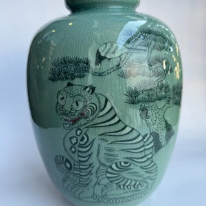 May include: A celadon green ceramic vase with a crackle glaze finish. The surface features a black ink illustration of a seated tiger with a red-accented mouth, positioned beneath a pine tree with a small bird perched on a branch.