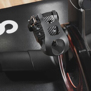 May include: Black exercise bike pedal with a black strap and silver buckle. The pedal has a cage-like design with a foot strap for secure use. The image shows a close-up of the pedal attached to the bike.