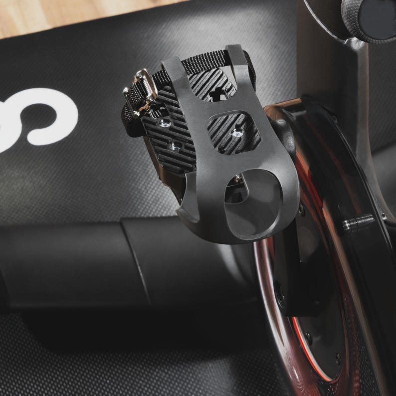 May include: Black exercise bike pedal with a black strap and silver buckle. The pedal has a cage-like design with a foot strap for secure use. The image shows a close-up of the pedal attached to the bike.