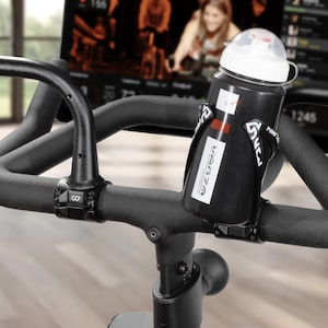 May include: Black water bottle with a white lid and a black holder attached to the handlebars of a stationary exercise bike. The bottle has a white label with the word "VANZA" printed on it. The bike's handlebars are black.