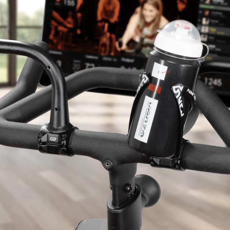 May include: Black water bottle with a white lid and a black holder attached to the handlebars of a stationary exercise bike. The bottle has a white label with the word "VANZA" printed on it. The bike's handlebars are black.