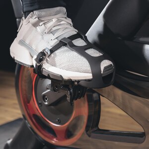 May include: Close-up of a gray athletic shoe secured to a stationary bike pedal with a black strap. The shoe has white stripes and a black cage-like cover over the top. The bike's flywheel is visible, with red accents.
