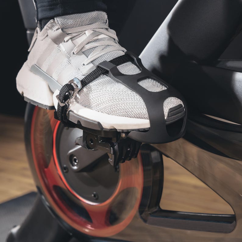 May include: Close-up of a gray athletic shoe secured to a stationary bike pedal with a black strap. The shoe has white stripes and a black cage-like cover over the top. The bike's flywheel is visible, with red accents.