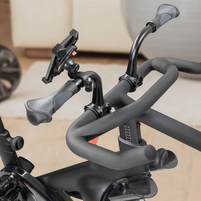 May include: Close-up of a black exercise bike handlebar with a phone holder attached. The handlebar has curved grips and a digital display. The phone holder is adjustable and holds a smartphone. The bike is designed for indoor cycling.