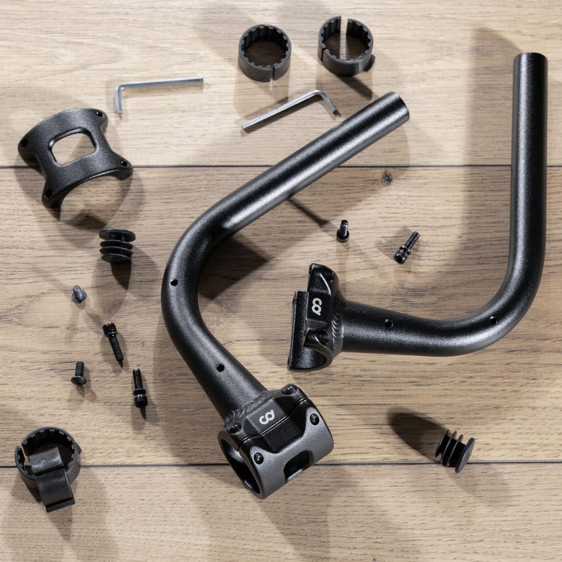 May include: An assortment of black bicycle handlebar components and hardware, including curved handlebars, mounting brackets, Allen wrenches, and small screws. The components are arranged on a light brown wooden surface.
