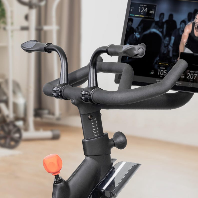 May include: Close-up of a black stationary exercise bike. The bike features curved handlebars with padded grips, a digital display showing workout metrics, and an orange adjustment knob. The bike is designed for indoor cycling workouts.