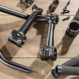 May include: An assortment of black bicycle components, including handlebars, stem, and grips, are arranged on a wooden surface. The components have a matte finish and are designed for a bicycle.