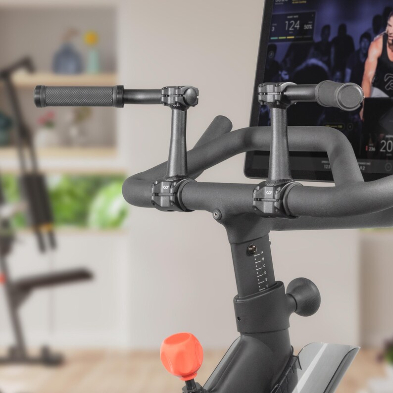 May include: Close-up of a stationary exercise bike. The bike features black handlebars with textured grips and a digital display. An orange adjustment knob is visible. The bike is designed for indoor cycling workouts.