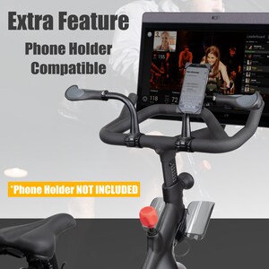 May include: Close-up of a stationary exercise bike with a phone holder. The bike is black with a red knob and a black seat. The text "Extra Feature Phone Holder Compatible" is visible, along with "Phone Holder NOT INCLUDED". A phone is mounted on the handlebars.